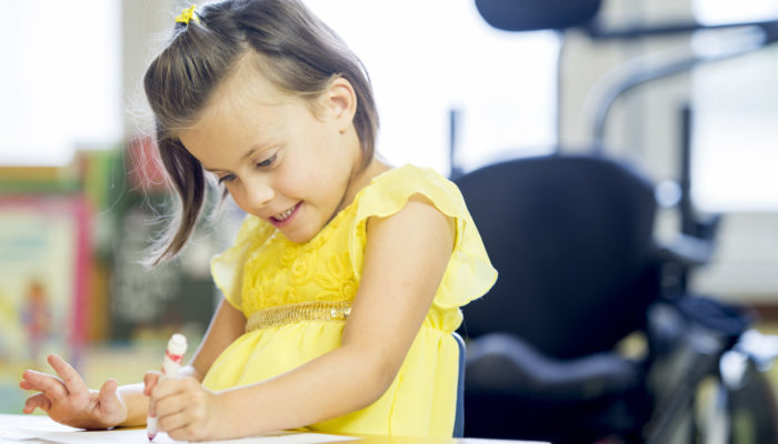 A happy little girl with cerebral palsy is sitting at a desk at an elementary school and is coloring a picture with a marker.
